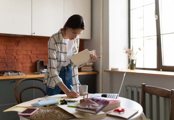 Focused young woman in shirt standing at table in kitchen and writing notes from textbook while studying at home