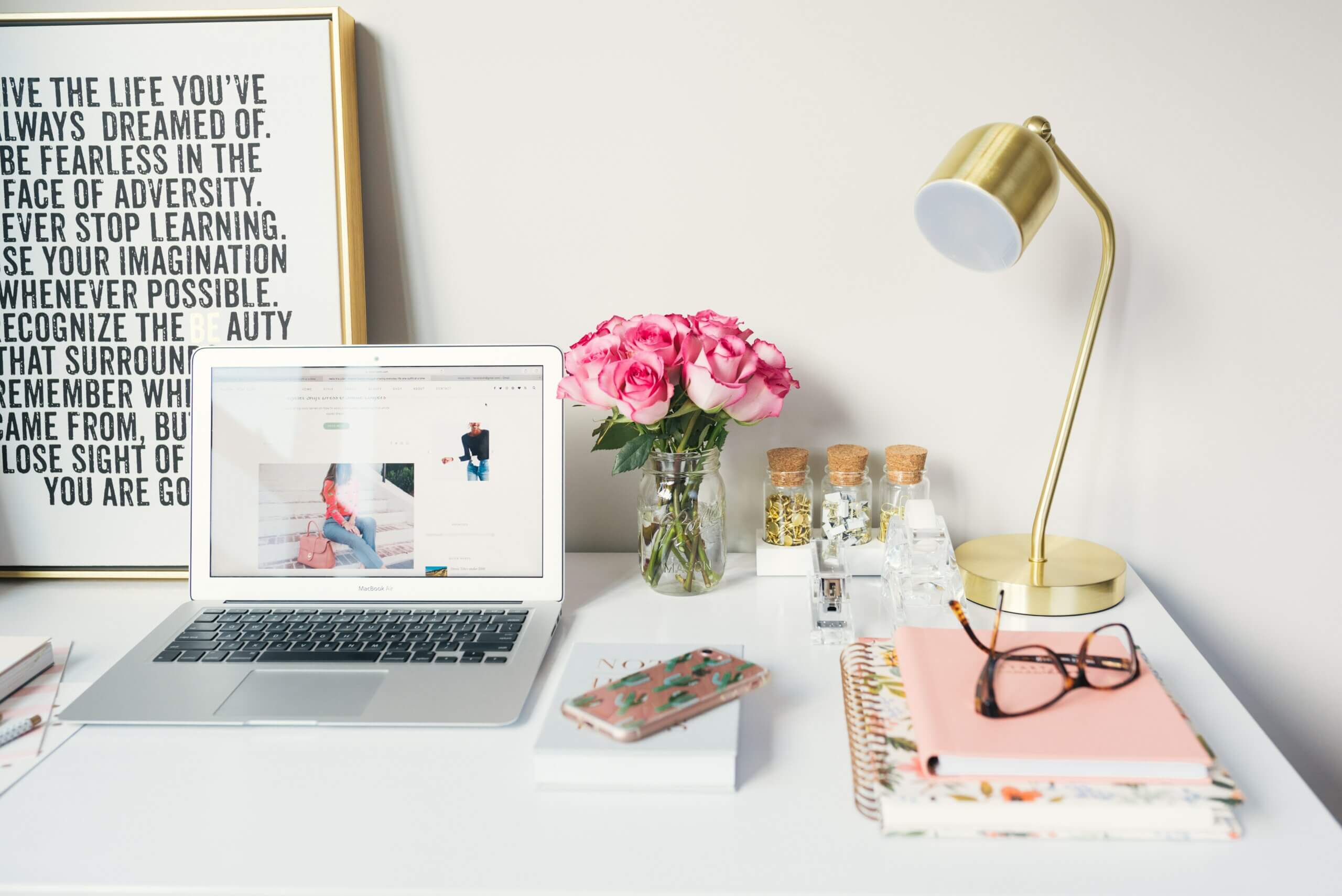 Desk with a macbook and books on top, glasses have been placed on the book and it has a golden lamp on their too.